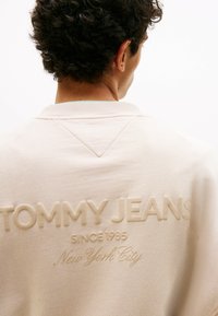 Hellbeige Sweatshirt mit geripptem Kragen, strukturiertem Stoff und geprägtem "TOMMY JEANS"-Logo auf der Rückseite mit Stadtdetails.