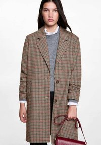 Rjav karirast plašč z dvorednim zapenjanjem, velikimi reverji in rjavimi gumbi. Nosi se ga preko sivega puloverja in drži rdečo torbico.