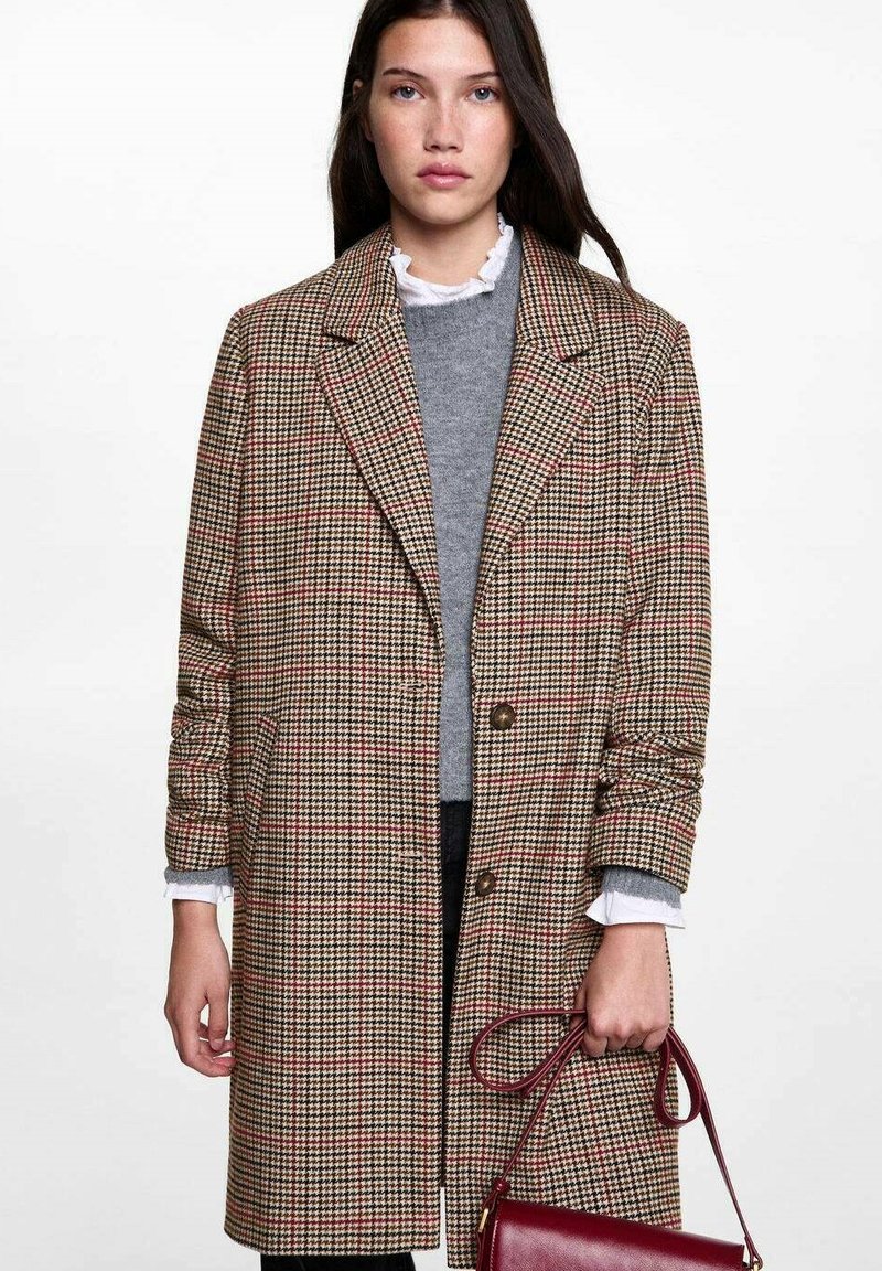 Rjav karirast plašč z dvorednim zapenjanjem, velikimi reverji in rjavimi gumbi. Nosi se ga preko sivega puloverja in drži rdečo torbico.