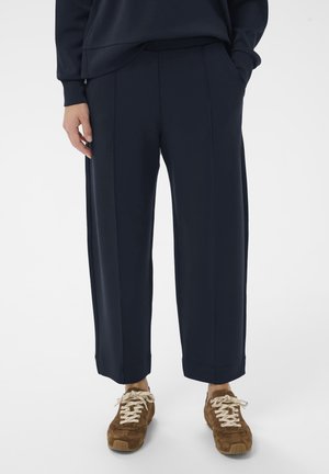 Personne portant un pantalon large foncé et des baskets marron à lacets, debout les mains dans les poches.