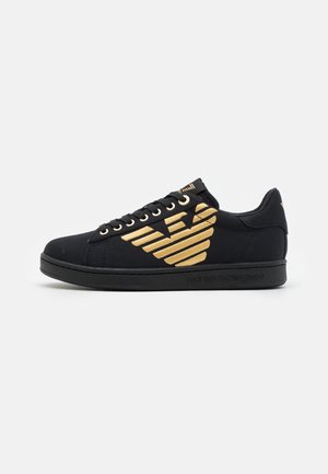 Baskets basses noires avec logo aigle doré sur le côté, semelle noire et lacets noirs sur fond uni.