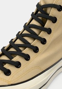 Baskets en toile beige avec des lacets et des œillets noirs. Présente un dessus texturé et une semelle en caoutchouc blanche avec une bande noire.