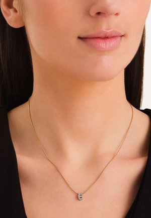 Złota naszyjnik z cienkim łańcuszkiem i okrągłym medalionem, zdobionym przezroczystymi kamieniami ułożonymi w dekoracyjny wzór.