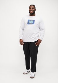 Grauer Sweatshirt mit blauem Grafiklogo auf der Vorderseite, kombiniert mit schwarzen Hosen und weißen Sneakern mit schwarzen Streifen. Das Model steht.