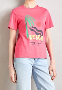 Camiseta de algodón rosa con un gráfico de una palmera, olas y la palabra "PLAYA" en amarillo. Manga corta y escote redondeado.