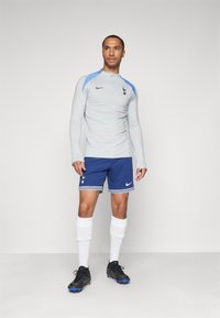 Hellgraues langärmliges Shirt mit blauen Akzenten, versehen mit einem Nike-Logo und dem Wappen von Tottenham; kombiniert mit blauen Shorts und schwarzen Fußballschuhen.