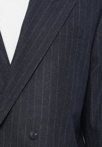 Blazer bleu marine à rayures avec un tissu texturé. Présente un design croisé, un unique bouton noir et un col en pointe.