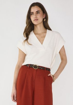 Woman with long brown hair wears a white V-neck short-sleeve top, rust-colored high-waist pants, braided brown belt, gold hoop earrings, and bracelet.