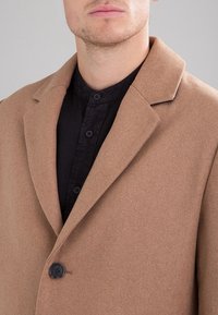 Homme vêtu d'un manteau en laine couleur camel par-dessus une chemise noire à boutons avec col mandarin, montré des épaules aux lèvres.