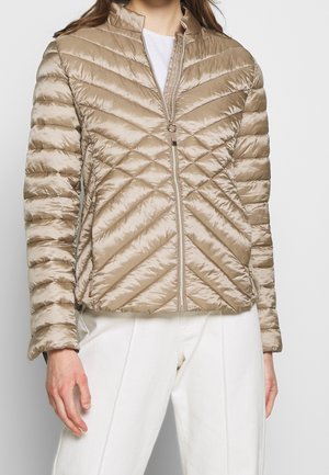 Femme portant une veste matelassée beige zippée avec des coutures diagonales et un pantalon blanc, debout devant un fond uni.