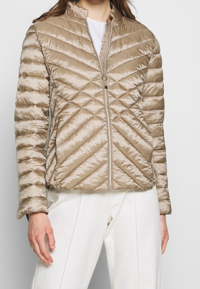 Femme portant une veste matelassée beige zippée avec des coutures diagonales et un pantalon blanc, debout devant un fond uni.