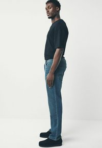 Melnā T-kreklā, zilās džinsu biksēs, pieguļošs, strukturēts audums, sānu skats, savienots ar melnajām slīdēšanas apaviem. Minimalistisks dizains, vienkāršas krāsas.