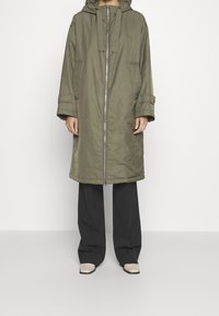 Cappotto lungo di colore verde oliva, con cappuccio, zip completa e polsini elastici. Abbinato a pantaloni neri a gamba larga e scarpe beige. Tessuto opaco, senza fantasie.