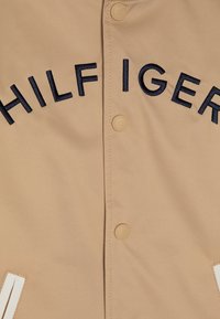 Giubbotto a vento beige con ricamo "HILFIGER" in blu navy. Bottoni con una finitura liscia e tasche laterali con strisce bianche. Tessuto resistente.