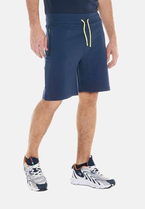 Homme portant un short bleu marine avec des cordons jaunes, une poche zippée noire et des chaussures de sport blanches et bleu marine avec des accents orange.