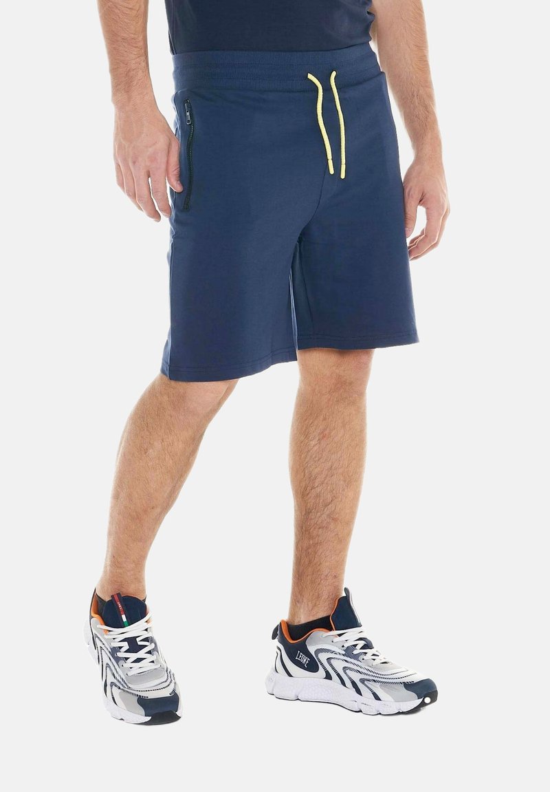 Homme portant un short bleu marine avec des cordons jaunes, une poche zippée noire et des chaussures de sport blanches et bleu marine avec des accents orange.