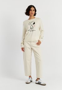 Maglione crema con grafica di Snoopy nera e dettagli rossi. Abbinato a pantaloni a gamba larga beige chiaro e sneakers bianche con strisce nere.