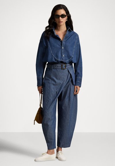 Sieviete valkā tumši zilu džinsa kreklu, jostu piegulošas plašas džinsa bikses ar augstu vidukli, baltas sporta kurpes, saulesbrilles un tur rokās mazu brūnu somiņu.