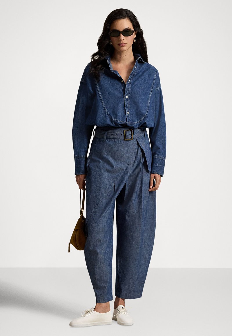 Donna con camicia di jeans scuro, pantaloni a gamba larga a vita alta con cintura, sneakers bianche, occhiali da sole e una piccola borsa marrone in mano.