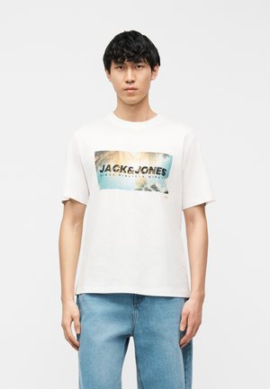 Giovane uomo in piedi, indossa una t-shirt bianca con la grafica di una palma e la scritta "JACK & JONES", abbinata a jeans azzurri.