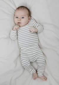 Prugasta bebi kombinacija u bijeloj i svijetlosivoj boji, s dugim rukavima i uskim krojem. Beba leži na mekoj, svijetloj podlozi.