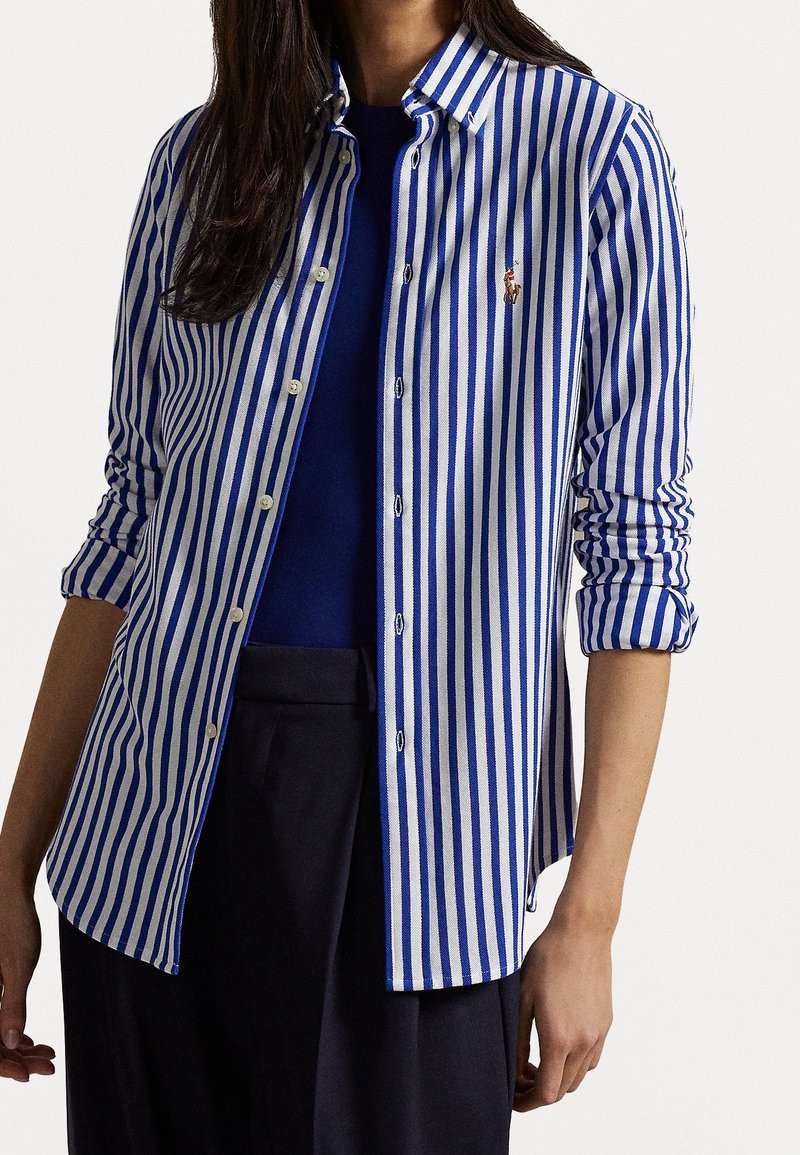 Femme portant une chemise à rayures verticales bleues et blanches boutonnée par-dessus un haut bleu foncé et un pantalon noir, manches retroussées.