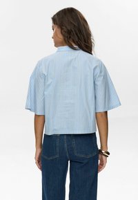 Lichte blauwe cropped shirt met verticale gele strepen, voorzien van korte mouwen en een staande kraag. Gekleed met een donkerblauwe jeans.