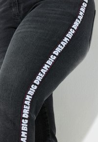 Jeans de denim negro con un atrevido texto en blanco y rosa "BIG DREAM" a lo largo de la costura lateral, con un diseño ajustado y tejido texturizado.