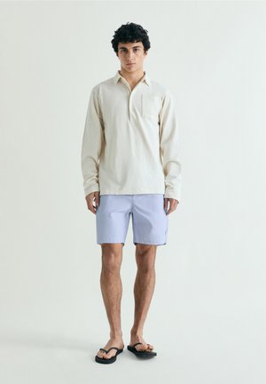 Joven de pie con una camisa polo de manga larga color crema, shorts azul claro y chanclas negras sobre un fondo liso.