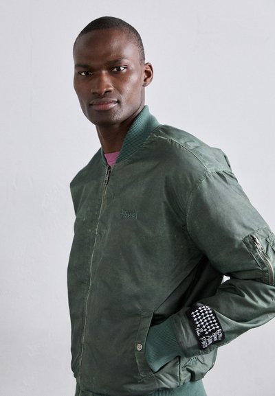 Veste bomber verte avec fermeture éclair à l'avant, poignets côtelés et poche latérale. Présente un tissu texturé et des accents à motifs sur la manche.