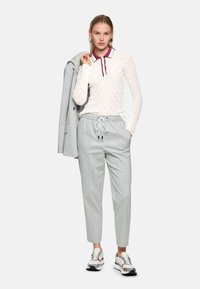 Weißes langärmeliges Oberteil mit strukturierten Mustern und burgunderrotem Kragen, kombiniert mit hellgrauen, gemusterten Hosen und weißen Sneakern mit Akzenten.