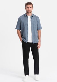 Chemise boutonnière à manches courtes bleu clair avec une poche poitrine, assortie à un pantalon noir et des baskets blanches sur un fond neutre.