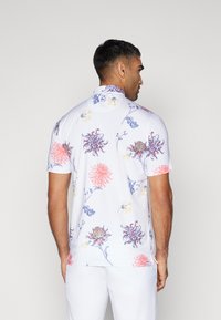 Kortermet skjorte i hvitt med et blomster mønster i rosa, blått og gult. Har kragehals og en avslappet passform bak.