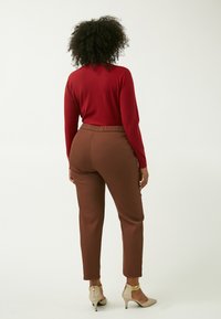 Rotes Langarmshirt, braune tapered Hose mit elastischem Bund und beige hohe Schuhe mit goldenen Akzenten. Glatter Stoff und figurbetontes Design.