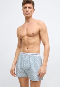 Άνδρας χωρίς πουκάμισο που φοράει λευκά και μπλε μποξεράκια με σχέδιο Calvin Klein, στέκεται μπροστά σε απλό φωτεινό φόντο.