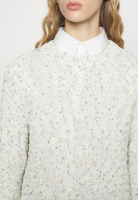 Strickpullover in hellem Creme mit einer strukturierten Musterung, die bunte Sprenkel in Lavendel und Gelb aufweist. Rundhalsausschnitt und lange Ärmel.