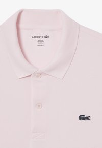 Ljusrosa polo skjorta gjord av en blandning av bomull och polyester. Har en klassisk krage, två knappar och en mörk krokodil-logotyp på vänster bröst.