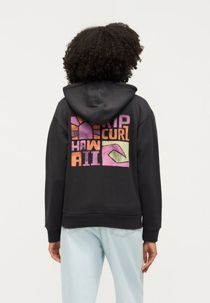 Person mit lockigem Haar, die einen schwarzen Hoodie mit buntem "Rip Curl Hawaii"-Motiv trägt und mit dem Rücken zu einem schlichten Hintergrund steht.