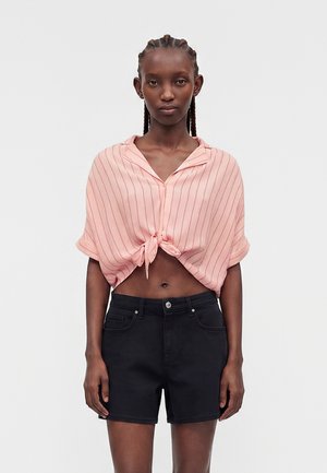 Kobieta z warkoczykami ma na sobie różowy, w paski, krótki rękaw, zawiązany w talii t-shirt i czarne, wysokie szorty na jednolitym tle.