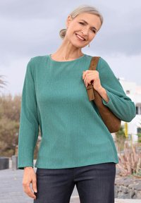 Lächelnde Frau mit grauem Haar in einem grünen strukturierten Langarmtop und dunklen Jeans, die an einem bewölkten Tag draußen eine braune Schultertasche hält.