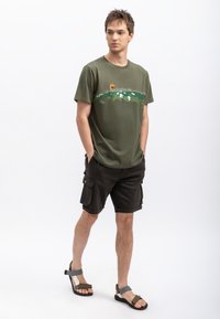 Olijfgroene katoenen t-shirt met een landschapprint; gecombineerd met donkere cargoshorts met zijzakken; model draagt bruine en grijze sandalen.
