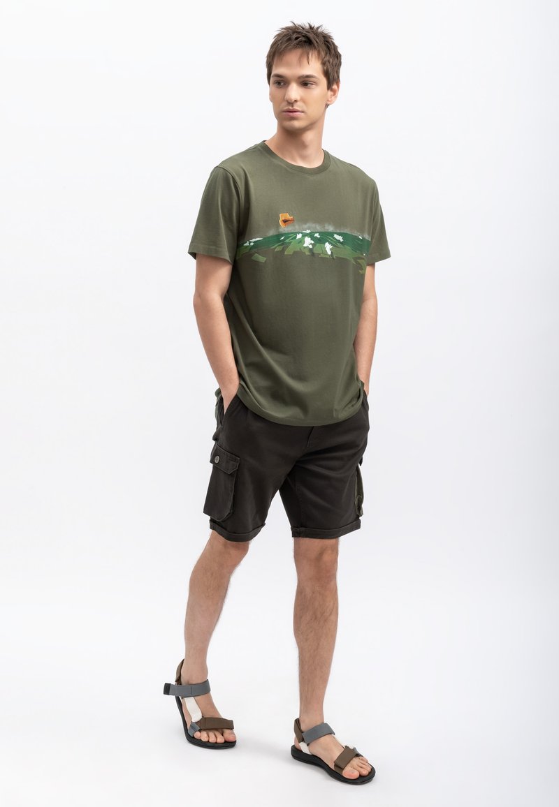 Olijfgroene katoenen t-shirt met een landschapprint; gecombineerd met donkere cargoshorts met zijzakken; model draagt bruine en grijze sandalen.