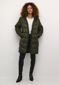Olive green puffer coat with a zip front, hood, and quilted design; paired with black jeans and ankle boots, set against a neutral background.