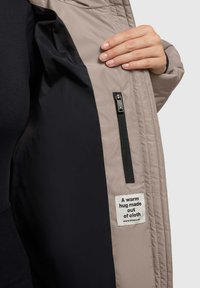 Beige quiltet jakke indvendigt med sort foring, der har en lynlåslomme og et mærke med teksten "Et varmt kram lavet af stof."