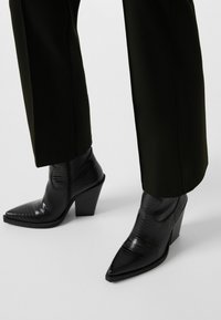 Svarta anklar stövlar med spetsig tå, tillverkade av strukturerat material, med en blockklack och subtila design detaljer. Kombinerade med mörka byxor.