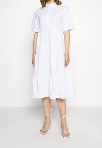 Abito camicia bianco in cotone con bottoni, maniche corte, design a strati e orlo che scende sotto il ginocchio, indossato con sandali bianchi con lacci.