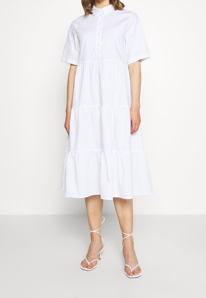 Abito camicia bianco in cotone con bottoni, maniche corte, design a strati e orlo che scende sotto il ginocchio, indossato con sandali bianchi con lacci.