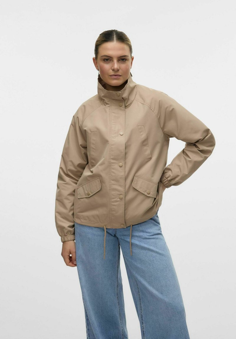 Beige vindjacka med tryckknappstängning, hög krage, elastiska ärmmuddar och två framfickor. Kombinerad med ljusblå vida jeans.