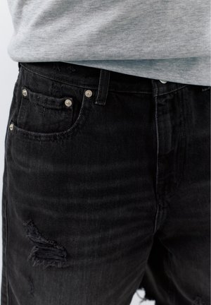 Gros plan sur un jean en denim noir usé avec des rivets visibles sur les poches et une chemise grise rentrée.