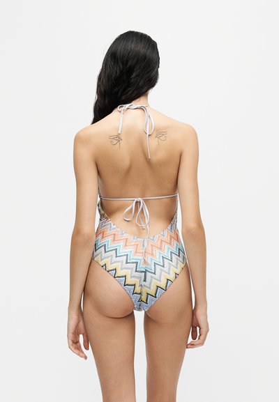 Femme aux longs cheveux foncés portant un maillot de bain une-pièce coloré à motif en zigzag, dos ouvert et bretelles nouées, montrant des tatouages sous les épaules.
